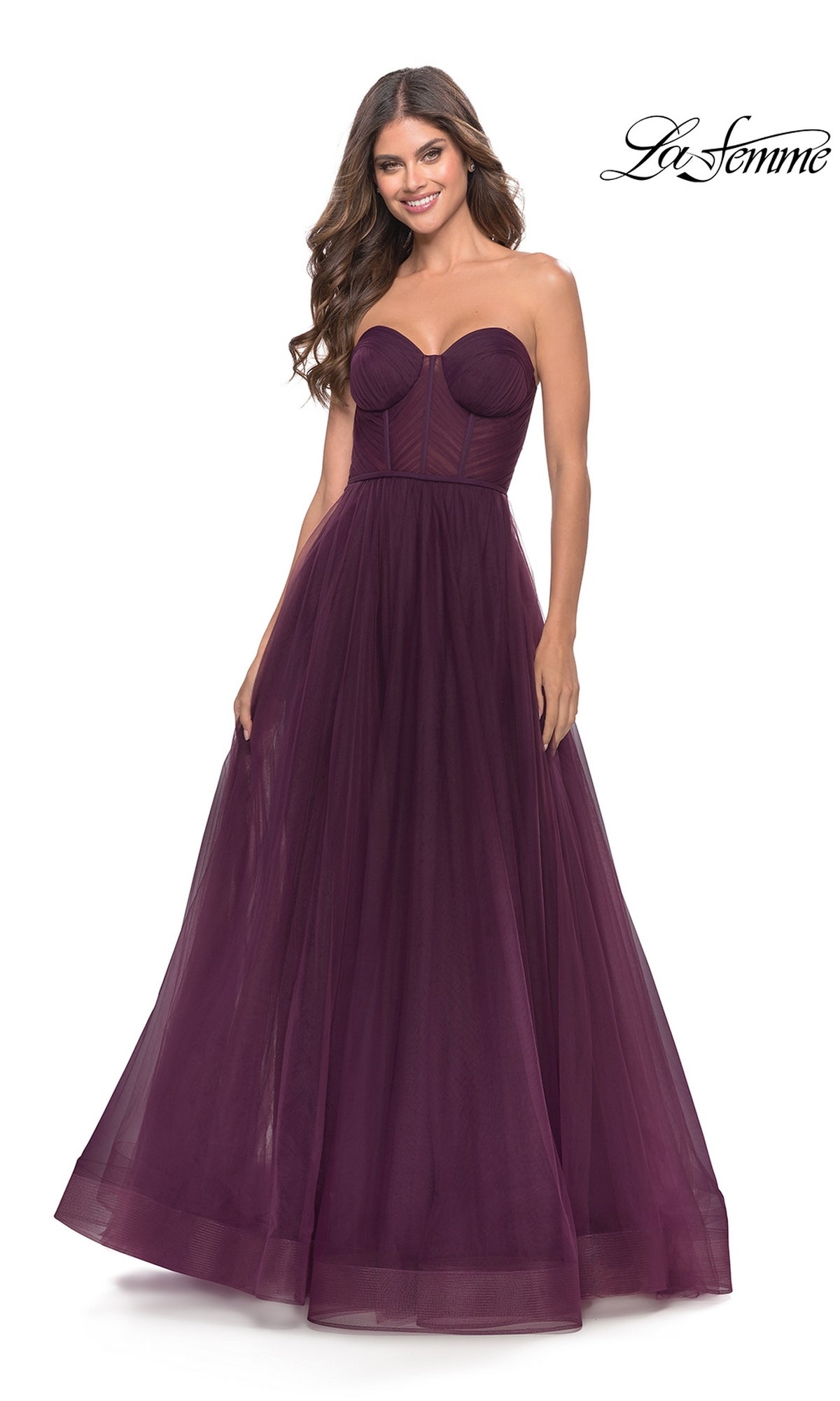 A woman stands smiling against a white background, wearing the Strapless Long Prom Ball Gown: La Femme 31205 in dark purple, featuring a fitted bodice and a tulle A-line ball gown skirt.